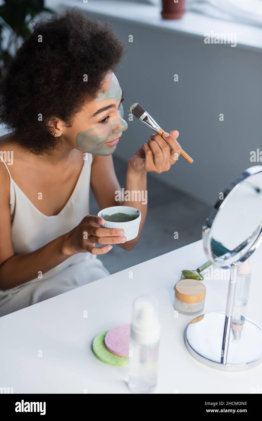 afroamerikanische Frau, die zu Hause eine Tonmaske neben kosmetischer Creme, Jadewalze und Spiegel anwendet Stockfoto