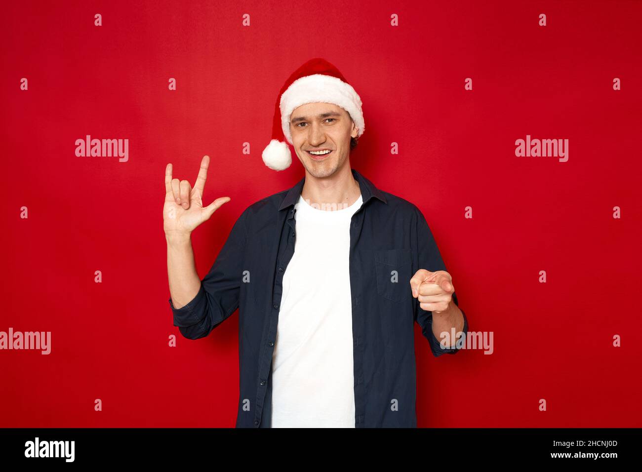 Lächelnder glücklicher junger Mann in lässiger Kleidung mit Neujahrshut zeigt Zeichen von taub und stumm mit seiner Hand Ich liebe dich isoliert auf rotem Hintergrund Raum für Textkonzept - Gesundheit, Kommunikation, Sympathie. Hochwertige Fotos Stockfoto