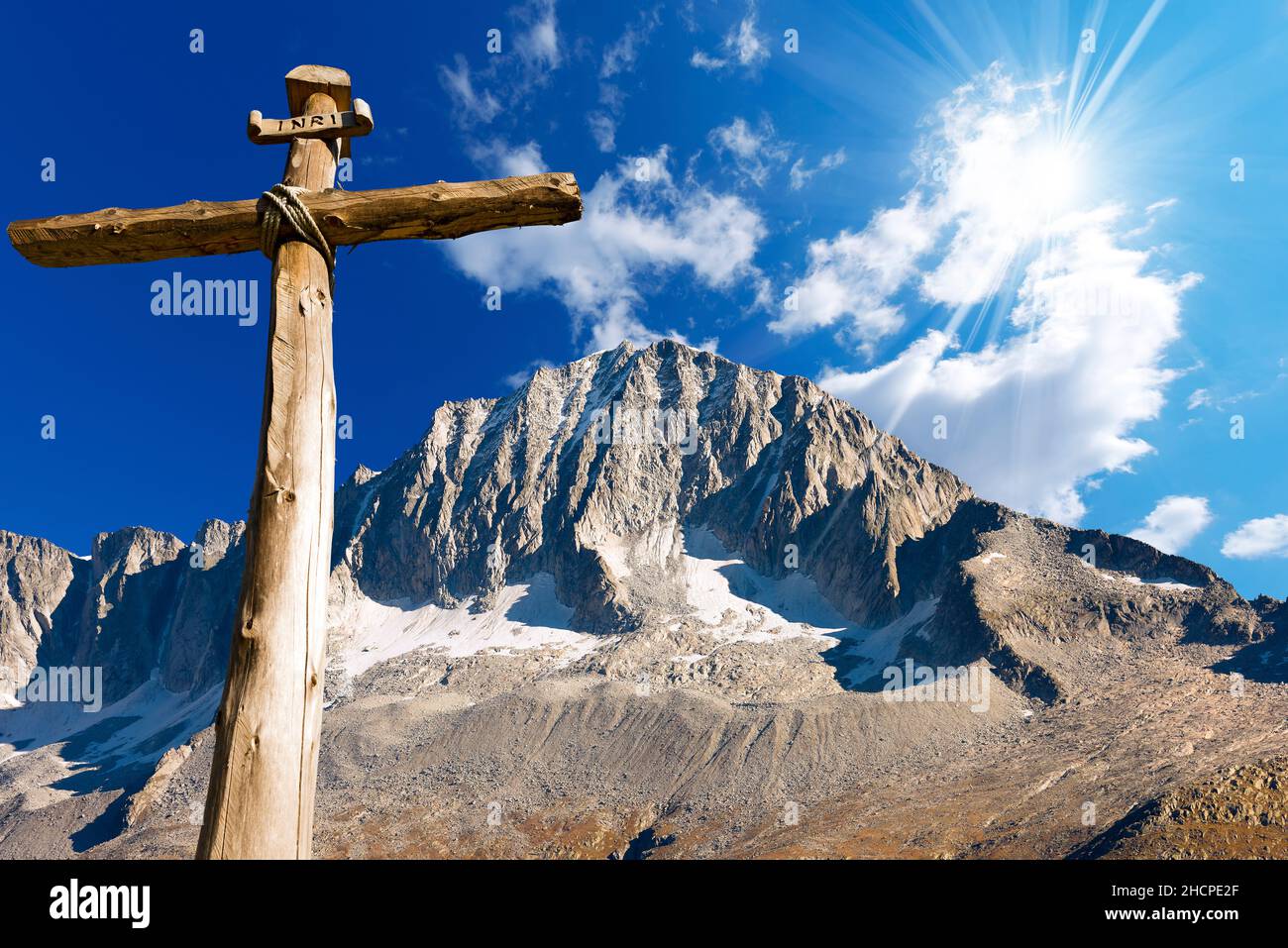 Altes Holzkreuz (Baumstämme) mit Seilen mit blauem Himmel, Wolken und Sonnenstrahlen gebunden. Nationalpark Adamello Brenta. Trentino Alto Adige, Italien. Stockfoto