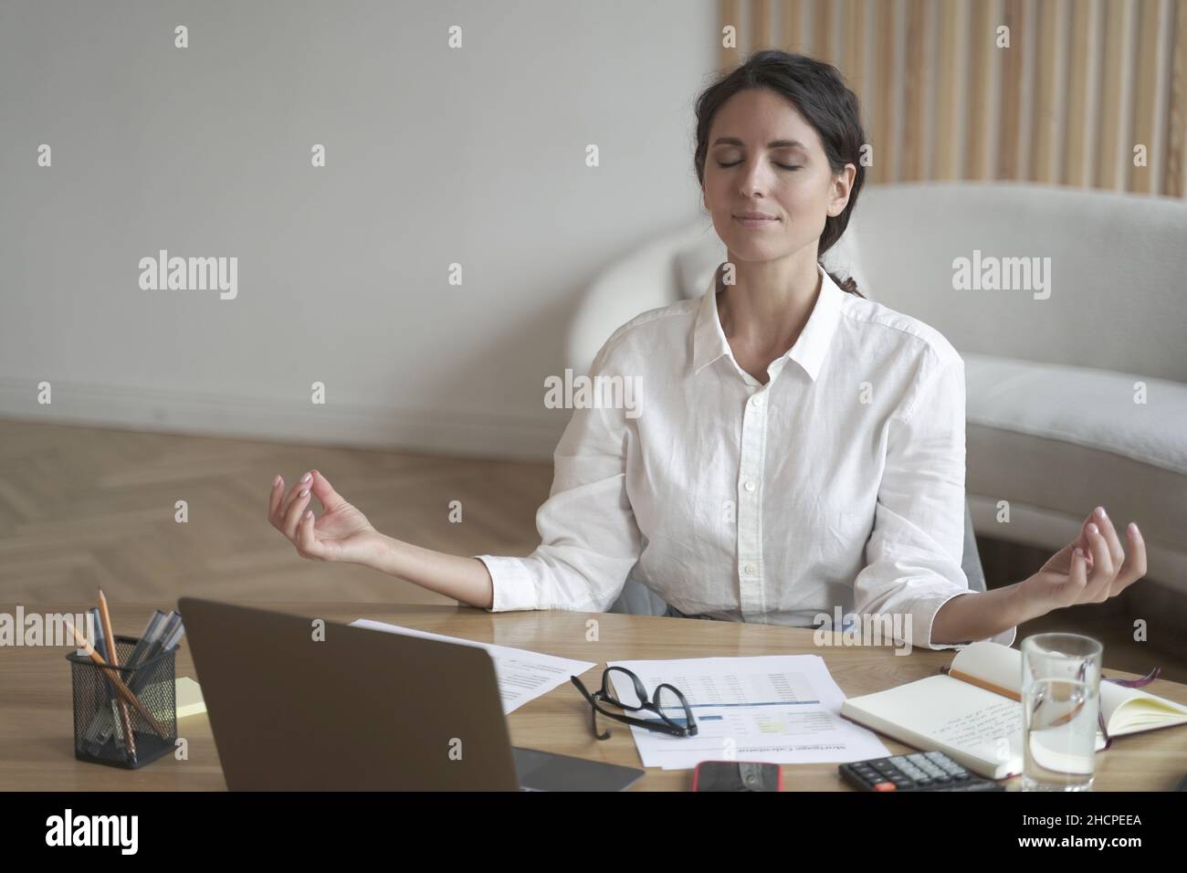 Ruhige Italienerin mit geschlossenen Augen meditiert am Arbeitsplatz, während sie mit einem Laptop am Tisch sitzt Stockfoto
