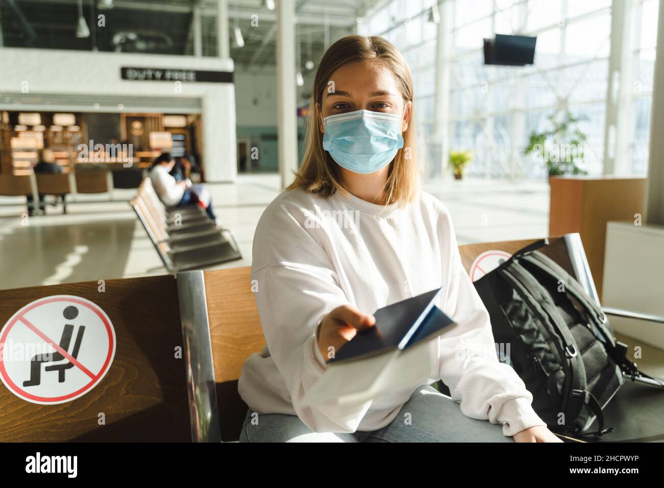 Junge Frau Reisende in medizinischer Maske sitzt im Wartezimmer und hält Pass und Ticket. Reisekonzept während der COVID 19-Epidemie Stockfoto