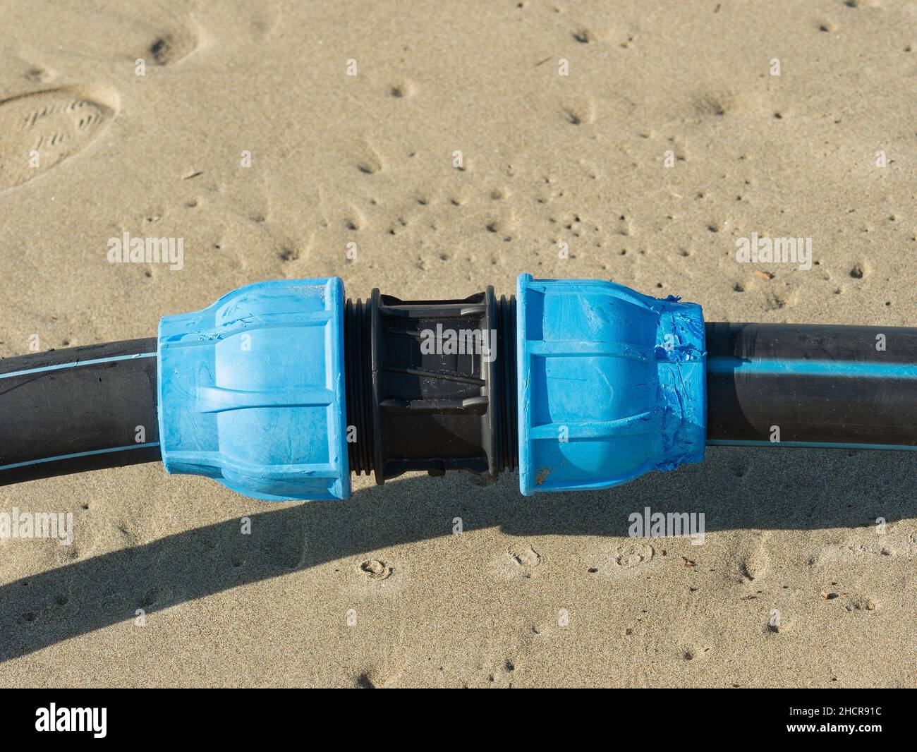 Mechanische Pressung aus Kunststoff, die zur Verbindung von zwei Polyethylenrohren für die Wasserversorgung verwendet wird. Schatten auf dem Sand im Hintergrund Stockfoto