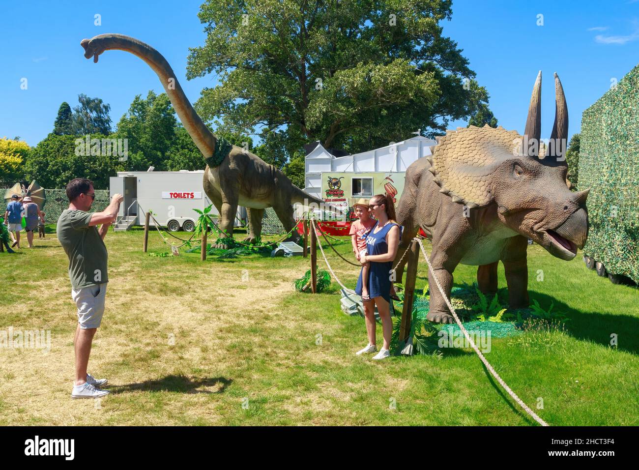 Eine Familie posiert bei einer Dinosaurier-Show für Fotos, mit den Dinosauriern Triceratops und Brachiosaurus im Hintergrund Stockfoto