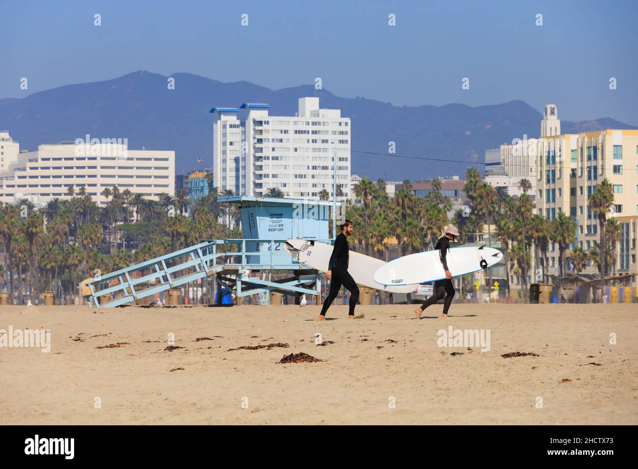 Surfer laufen den Strand vor einer Rettungsschwimmerhütte in Santa Monica, Kalifornien, USA, hinauf. Hotels in der Innenstadt im Hintergrund Stockfoto