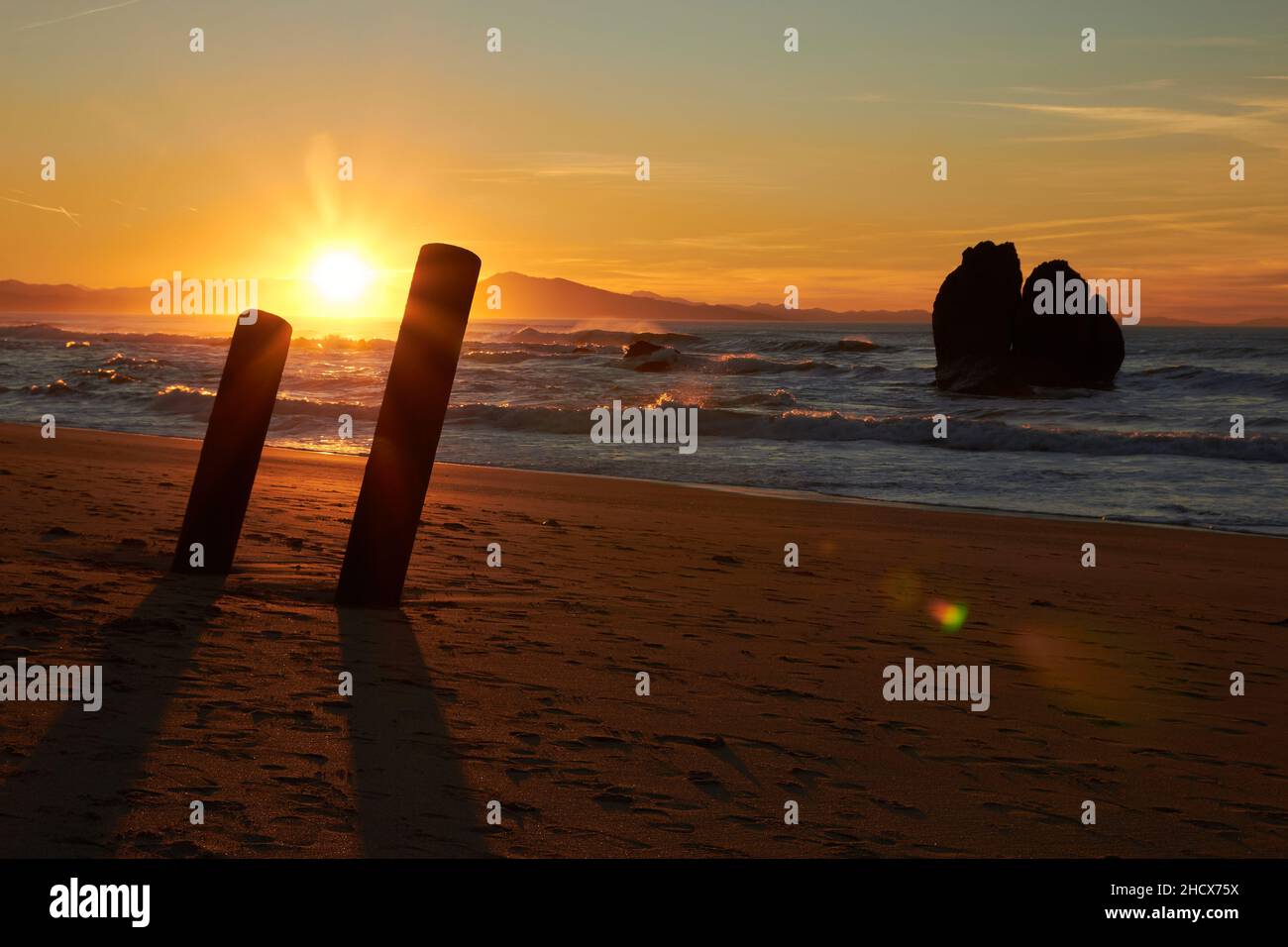 Landschaft eines Sonnenuntergangs, die Sonne versteckt sich im Hintergrund über dem Meer und hinterlässt warme Orange-Töne auf dem Sand des Strandes, mehrere Felsen schaffen eine Idylle Stockfoto