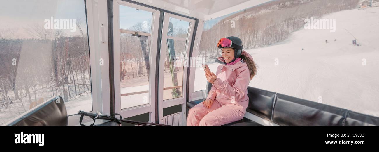 Ski Winter Holidays - Skifahrer mit Telefon-App in Gondelbahn. Lächelnde Frau, die auf das mobile Smartphone blickt, das Skibekleidung, Helm und Brille trägt Stockfoto