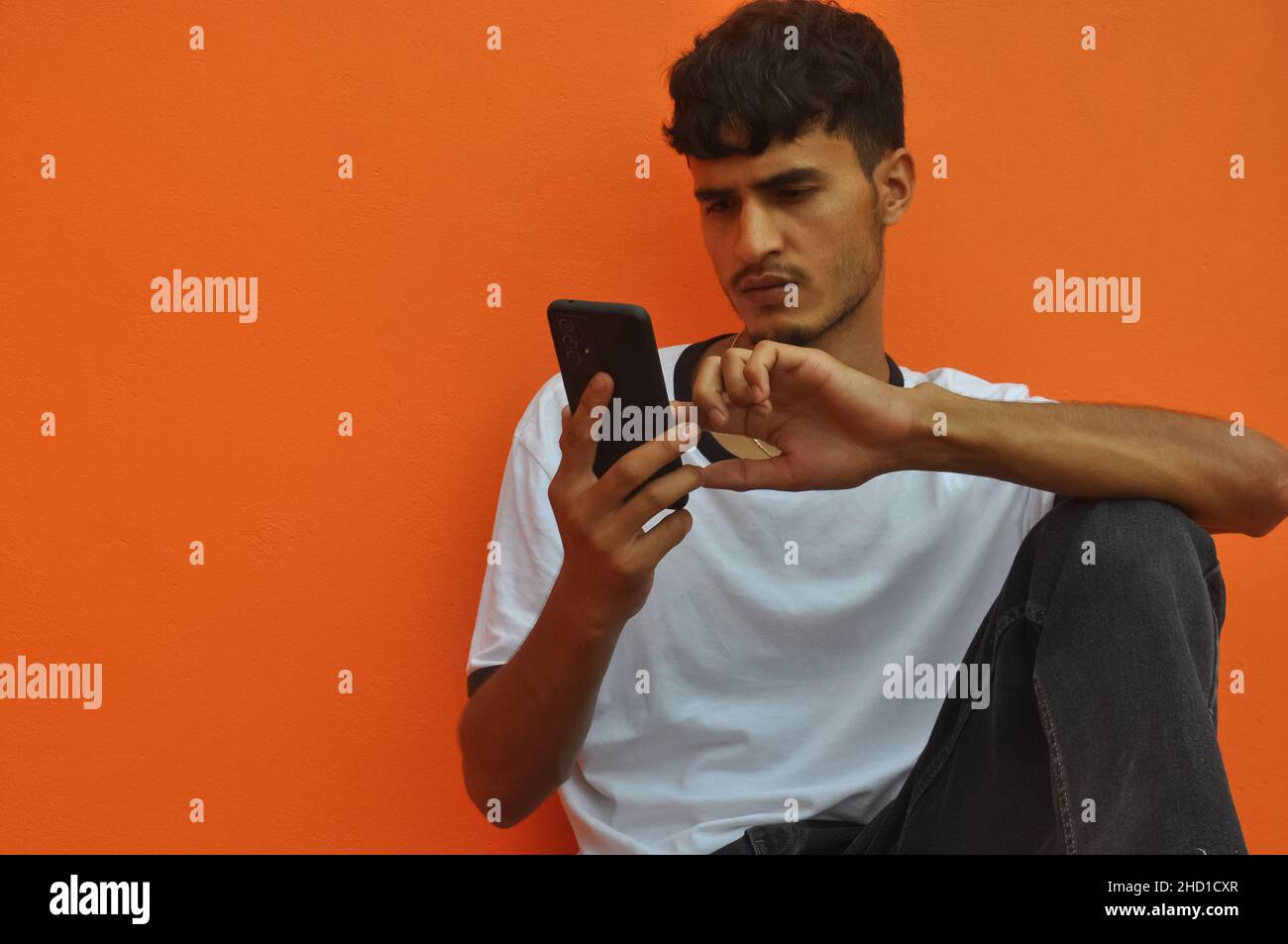 Ein hübscher Inder, der sein Telefon benutzt, während er auf einem orangen Wandhintergrund mit Kopierfläche sitzt Stockfoto