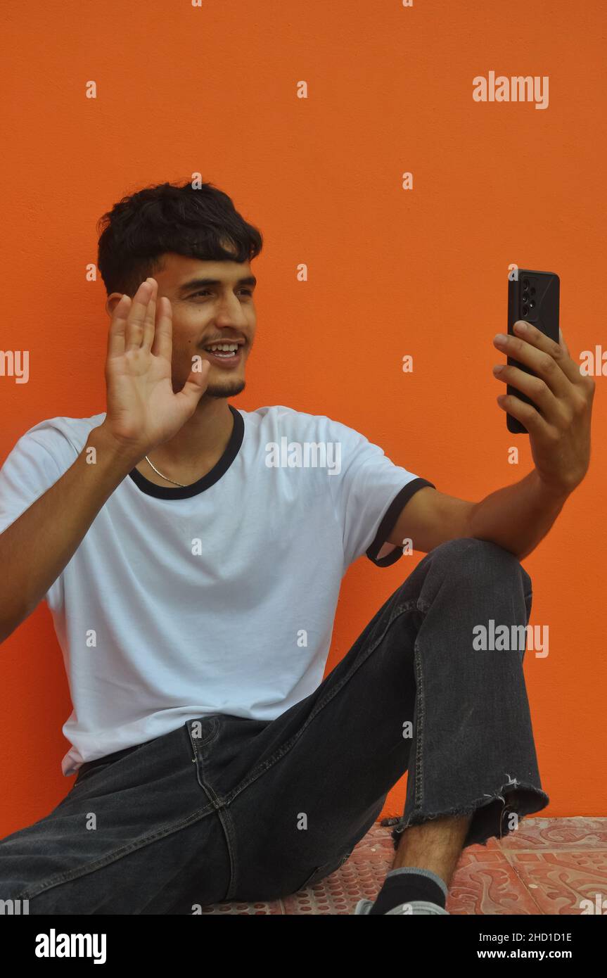 Ein fröhlicher junger Mann, der beim Videoanruf mit der Hand zu jemandem winkt und vor orangenen Wänden mit Platz zum Kopieren sitzt Stockfoto