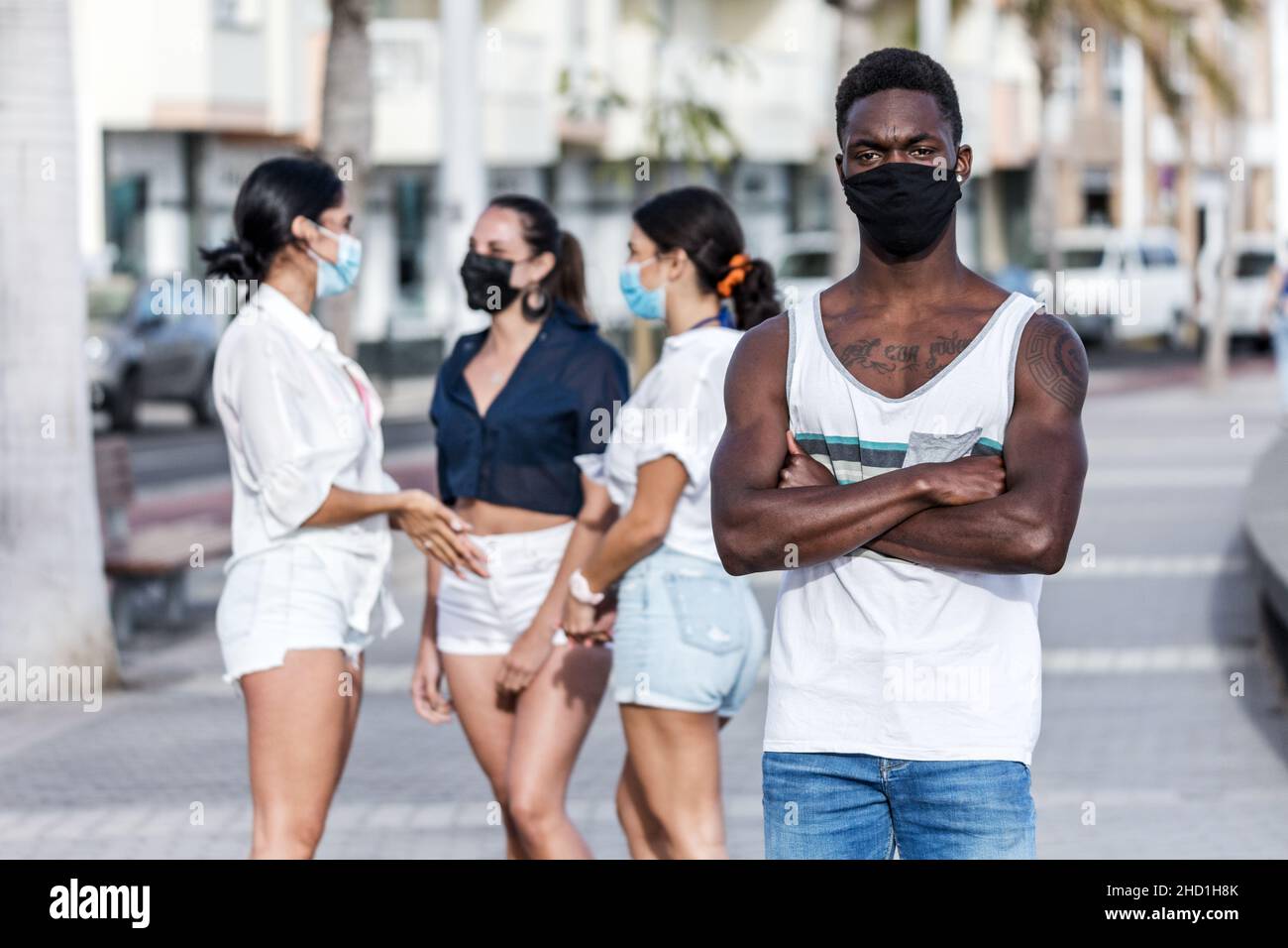 Düsteres afroamerikanisches Männchen in Maske, das mit gekreuzten Armen auf dem Hintergrund einer Gruppe weiblicher Freunde steht und sich einsam auf Lanzarote fühlt Stockfoto