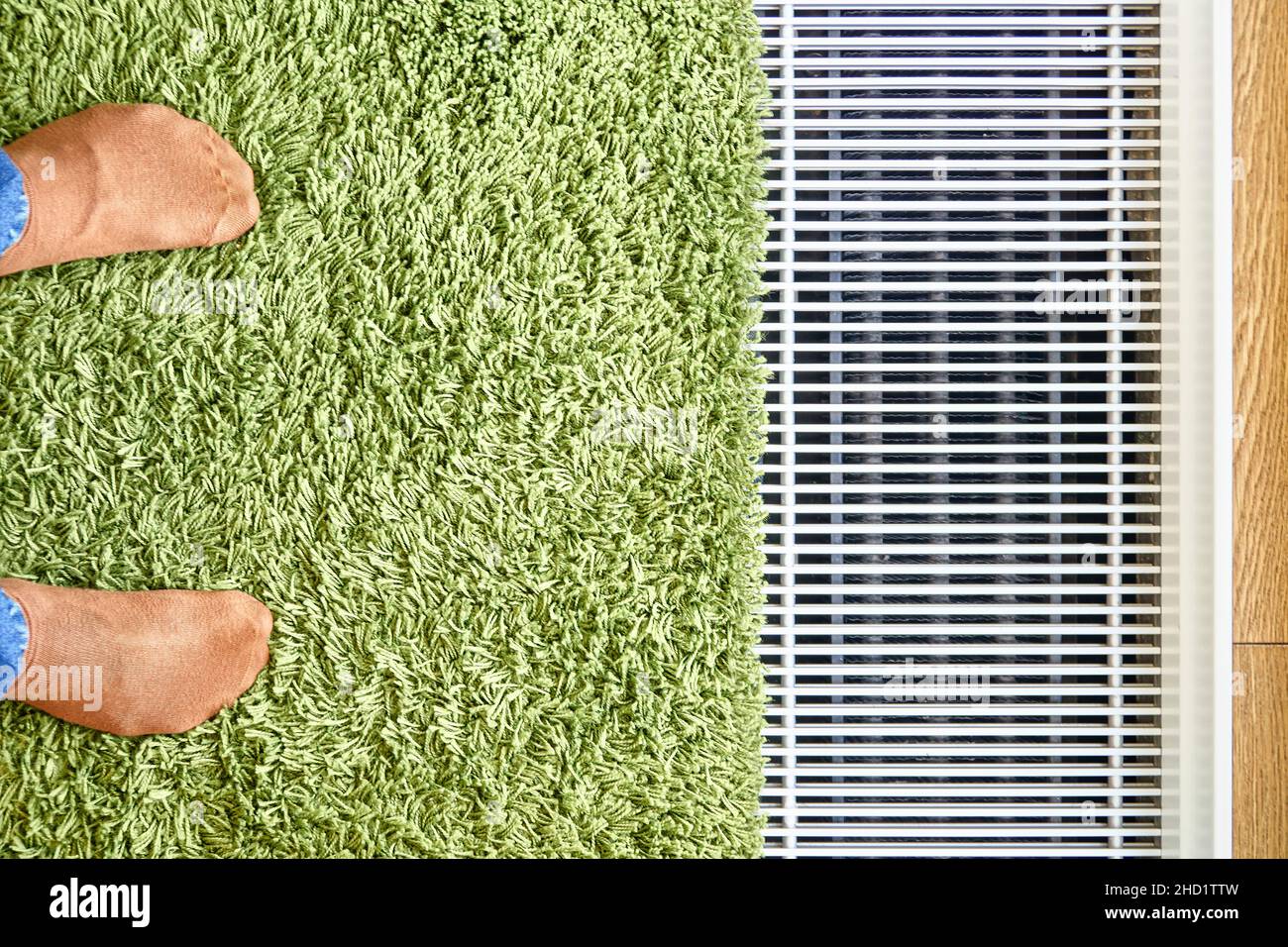 Moderner Wasser-Fußbodenwärmekonvektor in der Nähe des Panoramafensters mit grünem Teppich und Füßen in orangefarbenen Socken aus der oberen Ansicht installiert Stockfoto