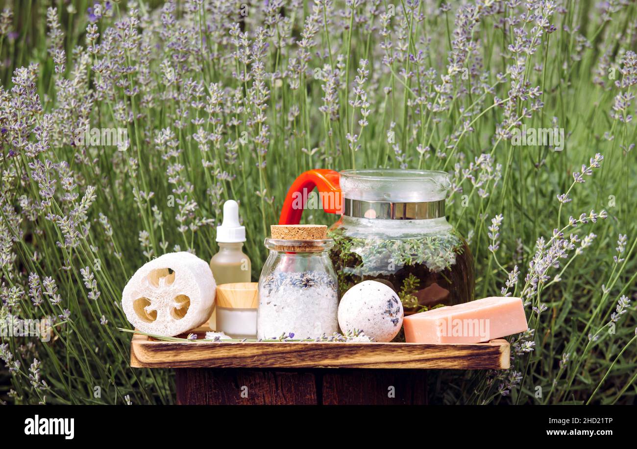 Verschiedene Lavendel-Spa-Produkte auf Holztablett im blühenden Lavendelfeld am sonnigen Sommertag. Glaskanne gefüllt mit heißem Lavendeltee-Getränk. Stockfoto