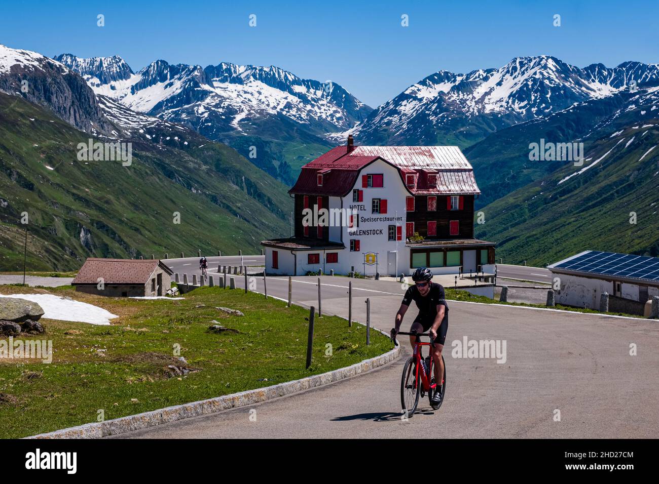 Ein Radfahrer, der die Furka Pass Straße hinauf fährt, vorbei am Hotel Galenstock, schneebedeckte Berge über Andermatt in der Ferne. Stockfoto