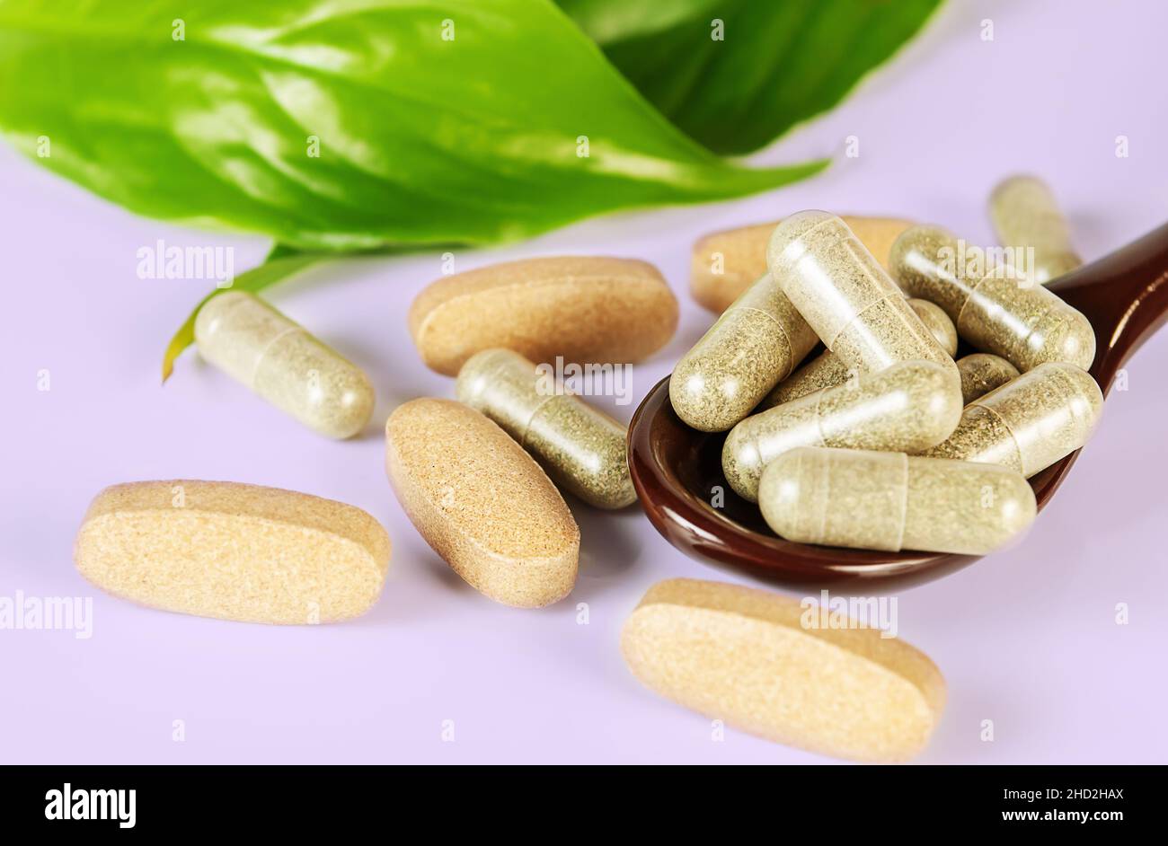 Brauner Löffel mit verschiedenen Arten von Kräutertabletten und Kapseln und grünen Blättern auf hellblauem Hintergrund aus nächster Nähe. Konzept der Kräutermedizin Stockfoto