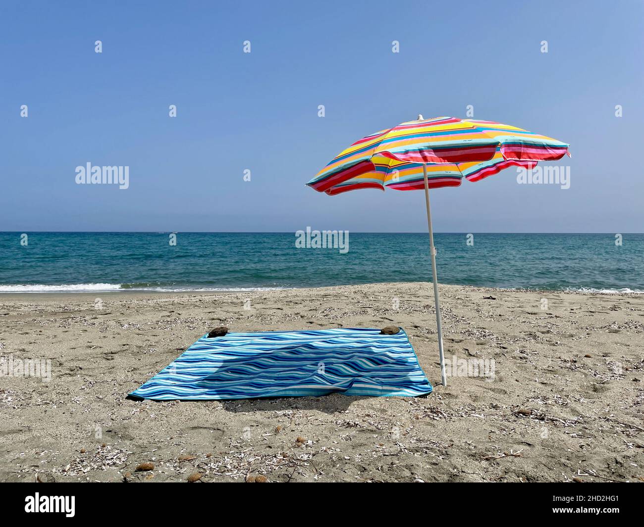 Farbenfroher Regenschirm und blaue Decke am einsamen Sandstrand vor blauem Himmel. Stockfoto