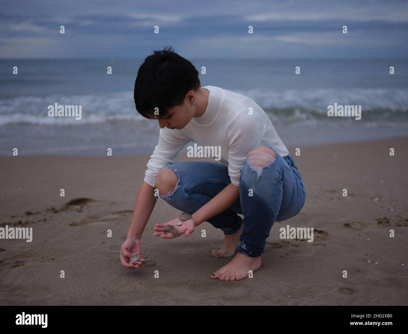 Nicht-binäre Frau, die Muscheln aus dem Sand am Strand aufholt Stockfoto