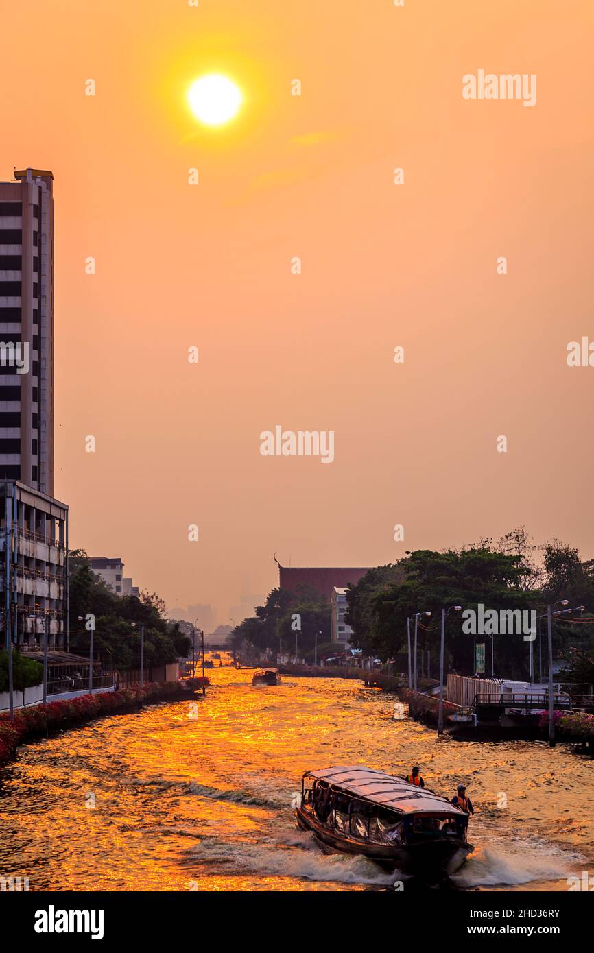 Wassertaxis kommen in einem Bereich, in dem Passagiere abgesetzt werden, in Bangkok an. Sie sind aufgereiht, aber aus der Ferne. Der Sonnenuntergang erzeugt ein goldenes Leuchten auf dem Wat Stockfoto