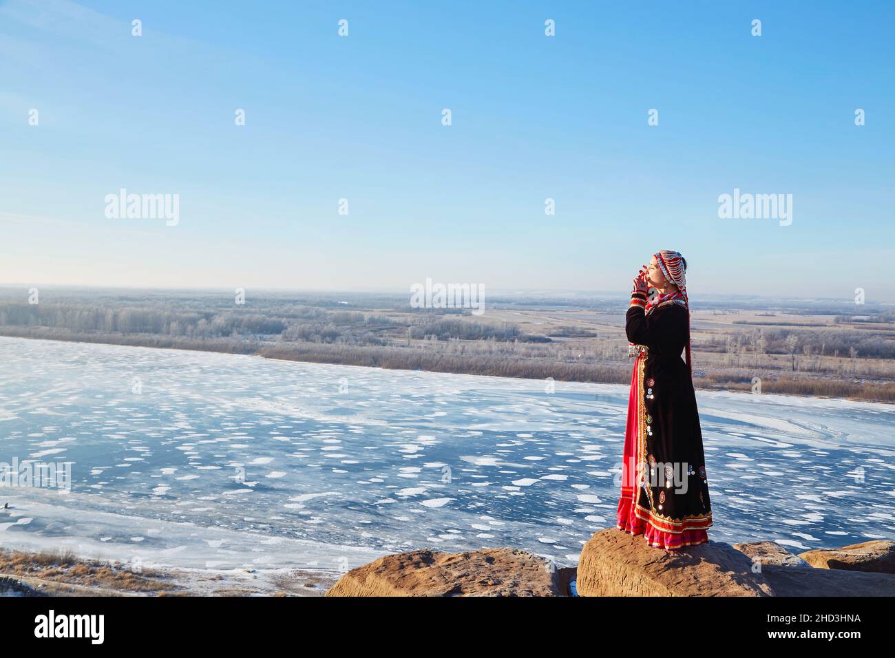 Seitenansicht der Frau in authentischem Kostüm spielt ein Musikinstrument maultrommel mit dem gefrorenen Fluss auf dem Hintergrund, während sie auf Steinen ne steht Stockfoto
