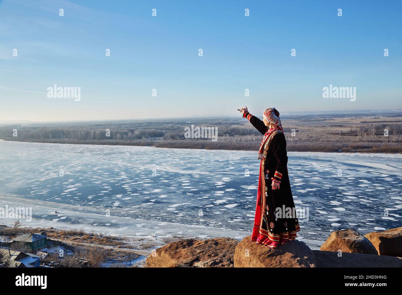 Seitenansicht einer Frau in authentischem Kostüm, die den Arm zum gefrorenen Fluss streckt, während sie an einem wolkenlosen Tag auf dem Land in der Nähe des Dorfes auf Steinen steht Stockfoto