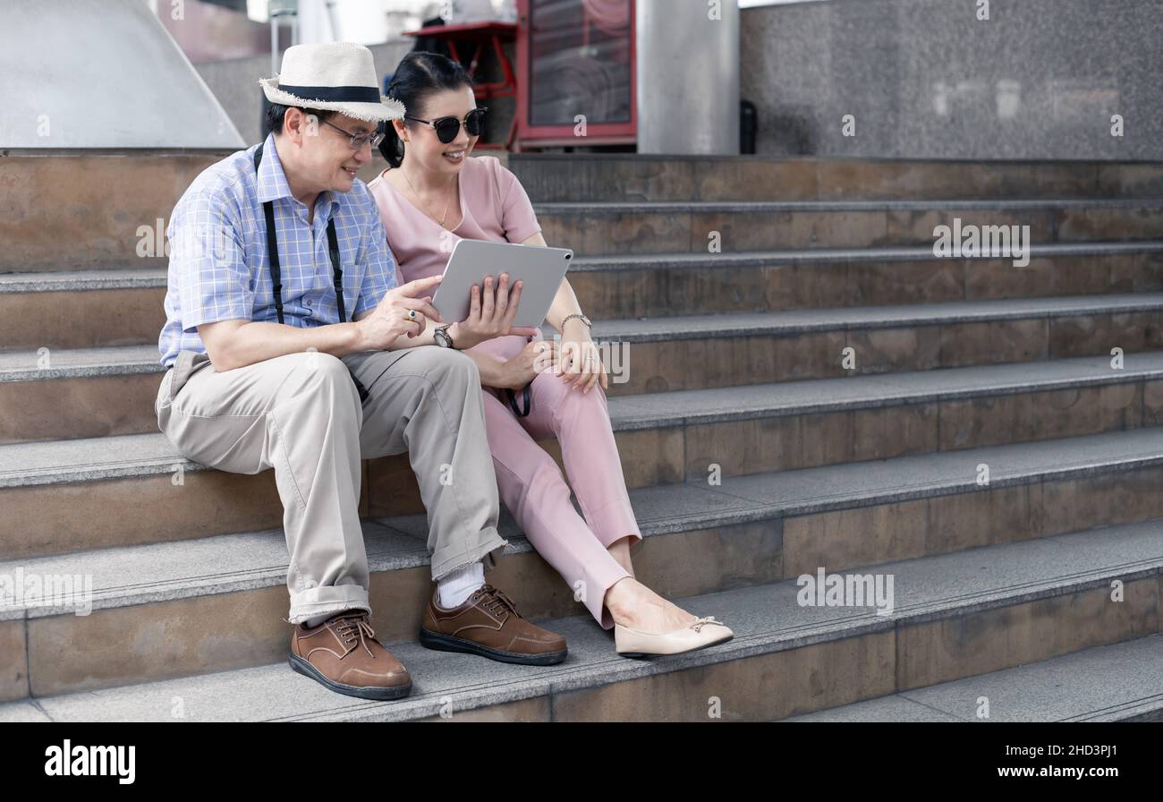 Ältere asiatische Paare sitzen auf der Treppe, planen, finden Reiseinformationen per Tablet mit einem Lächeln. Reisekonzept für ältere Paare. Tante und Onkel sitzen auf der Treppe Stockfoto