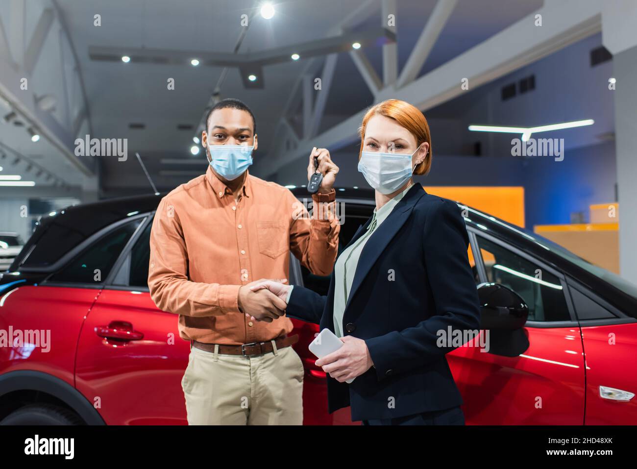 Autohändler und afroamerikanischer Kunde in medizinischen Masken, die sich beim Betrachen der Kamera die Hände schütteln Stockfoto