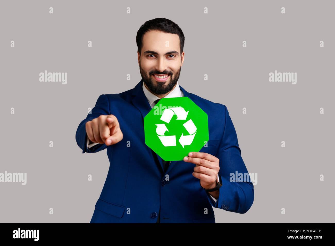 Positiver optimistischer Mann mit grünem Recycling-Schild, Umweltschutz, Ökologie-Konzept, zeigt auf Kamera, trägt offiziellen Stil Anzug. Innenaufnahme des Studios isoliert auf grauem Hintergrund. Stockfoto