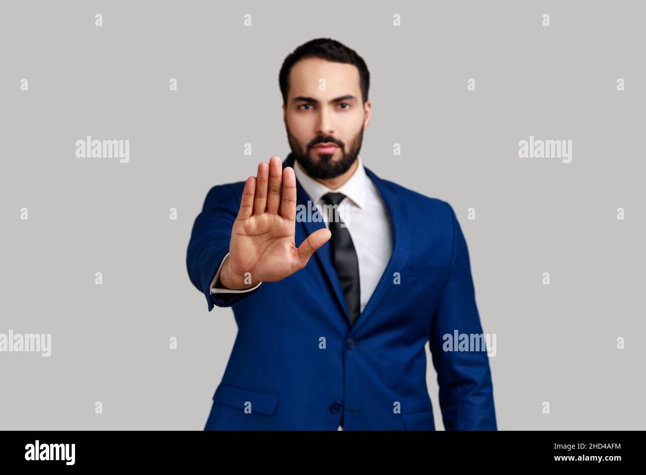 Ernsthafter bärtiger Mann, der eine Stop-Geste mit Handfläche macht, Konfliktverbotswarnung vor Gefahren, Mobbing aufhört und einen offiziellen Anzug trägt. Innenaufnahme des Studios isoliert auf grauem Hintergrund. Stockfoto