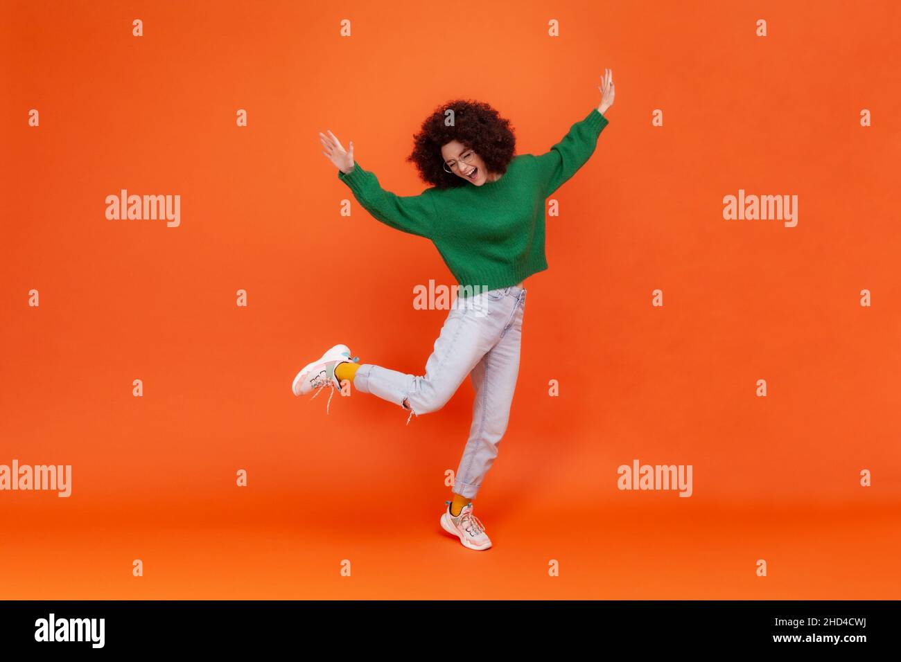 In voller Länge Porträt einer glücklichen Frau mit Afro-Frisur tragen grünen lässigen Stil Pullover auf einem Bein stehen, erhöhte Arme, tanzen, feiern. Innenaufnahme des Studios isoliert auf orangefarbenem Hintergrund. Stockfoto