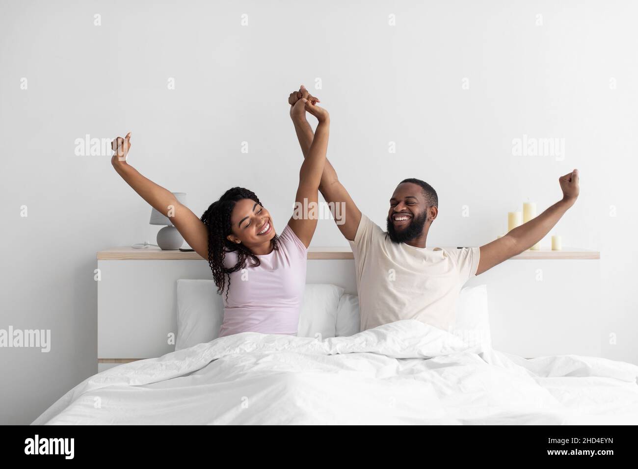 Zufriedener junger afroamerikanischer Kerl und Dame heben die Hände und strecken den Körper im Schlafzimmer auf dem Bett Stockfoto