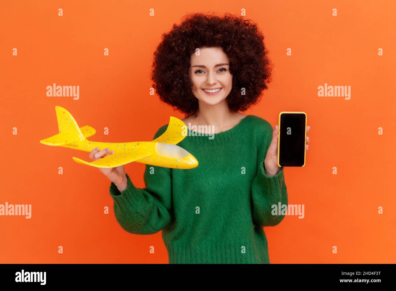 Lächelnde Frau mit Afro-Frisur trägt einen grünen Pullover mit gelbem Flugzeug und Smartphone mit schwarzem leerem Bildschirm, Kopierraum. Innenaufnahme des Studios isoliert auf orangefarbenem Hintergrund. Stockfoto