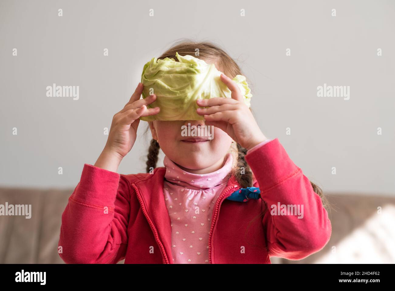 Das kleine Mädchen bedeckt ihr Gesicht mit einem Kohlblatt aus dem heimischen Garten. Psychologie. Kinderspiele. Gesunde Lebensmittel ohne GVO. Stockfoto