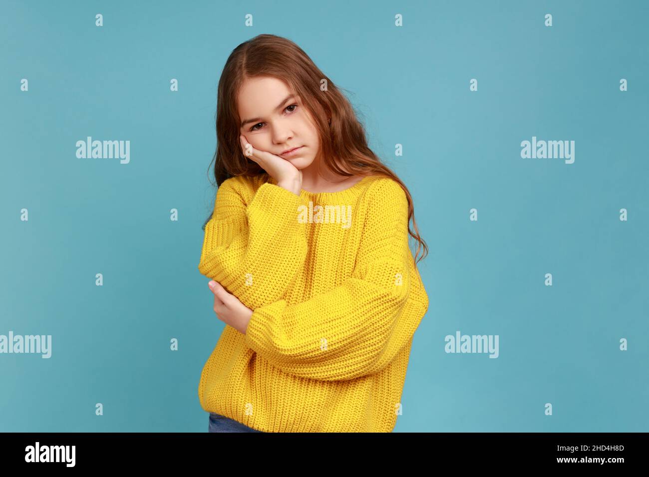 Kleines Mädchen lehnt sich an die Hand, sieht deprimiert aus, Kind mit gleichgültigem düsteren Ausdruck hat keine Energie, trägt gelben Pullover im lässigen Stil. Innenaufnahme des Studios isoliert auf blauem Hintergrund. Stockfoto