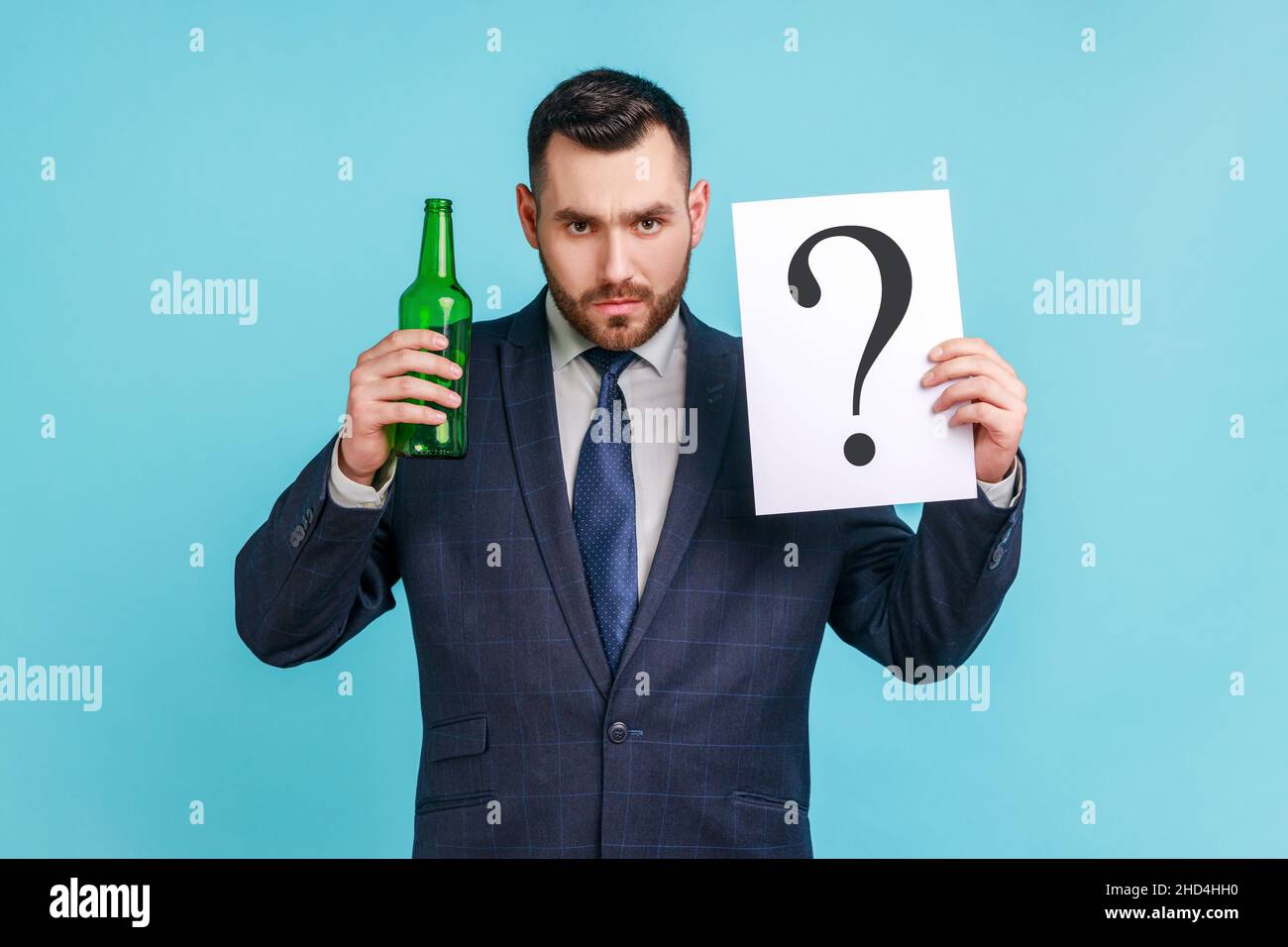 Trinken oder nicht. Porträt eines ernsthaften Mannes mit Bart, der einen dunklen Anzug im offiziellen Stil trägt, der eine Flasche mit Alkohol und Papier mit Fragezeichen hält. Innenaufnahme des Studios isoliert auf blauem Hintergrund. Stockfoto