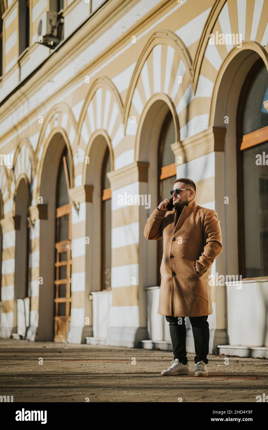 Aufnahme eines kaukasischen Mannes mit langem Mantel, der auf der Straße in Bosnien und Herzegowina telefoniert Stockfoto