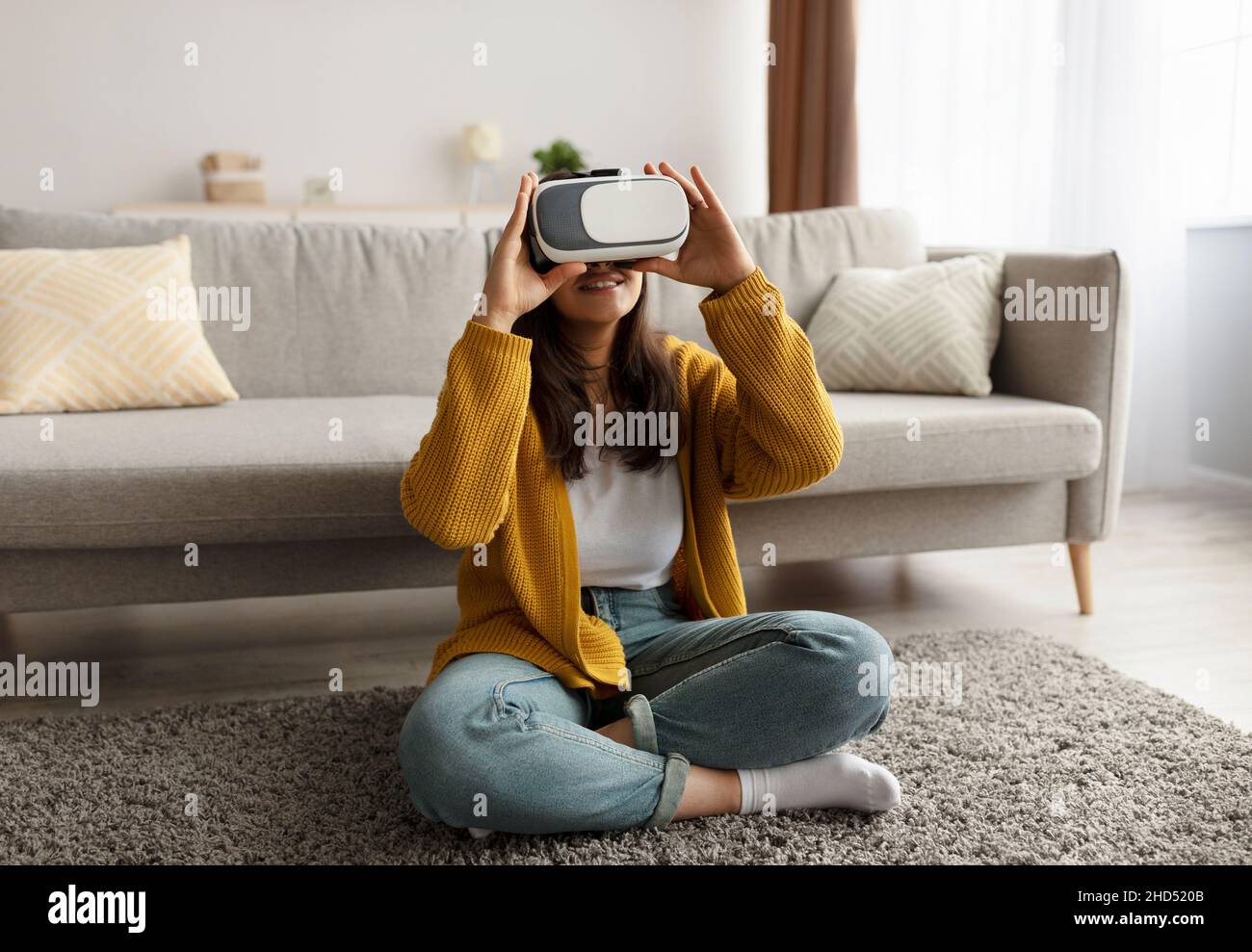 Junge arabische Frau in legerem Outfit mit moderner VR-Brille, während sie auf dem Boden sitzt und Virtual Reality-Session erlebt Stockfoto