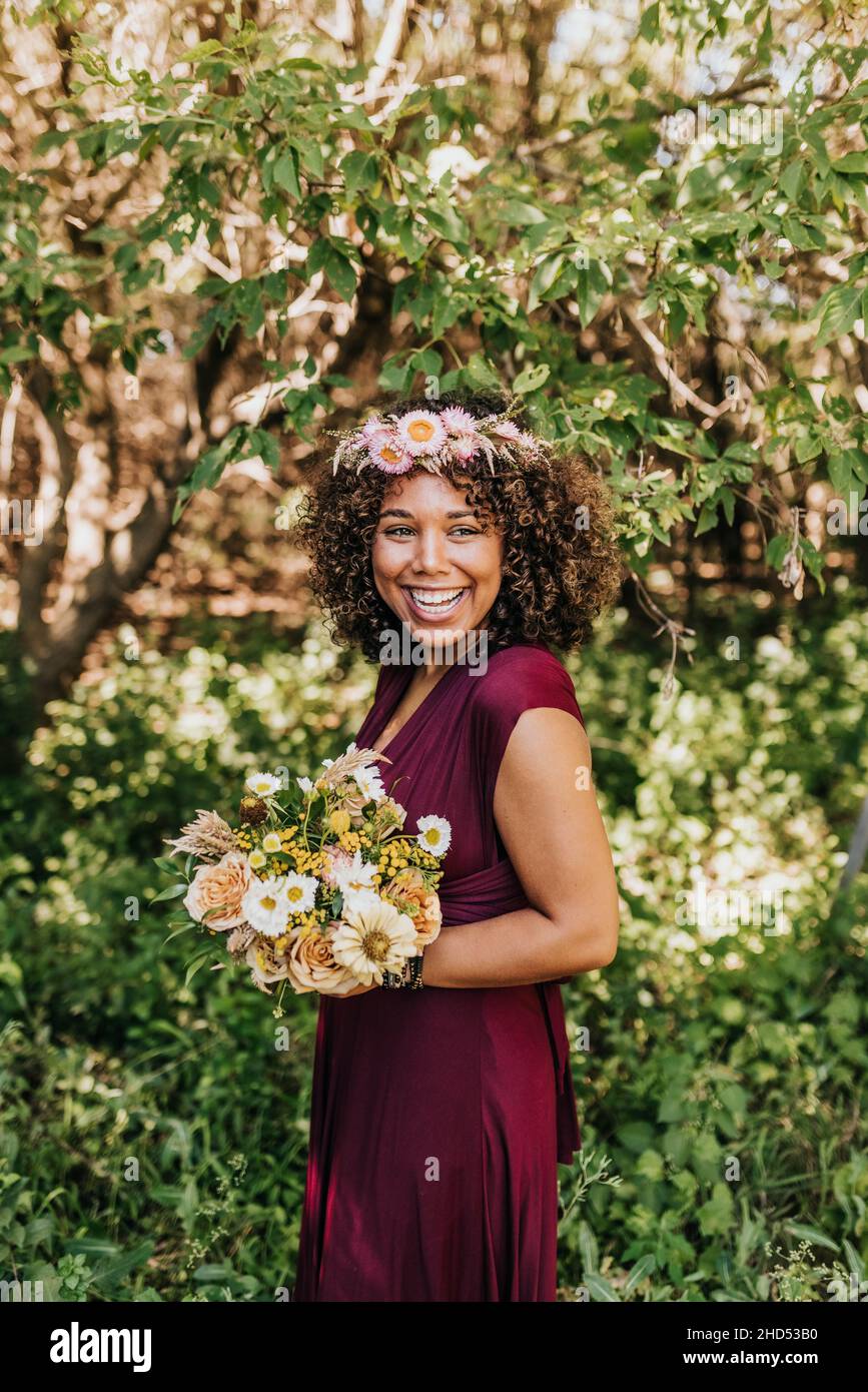 Lächelnde Frau steht im Wald mit Blumenstrauß und trägt die Blumenkrone Stockfoto