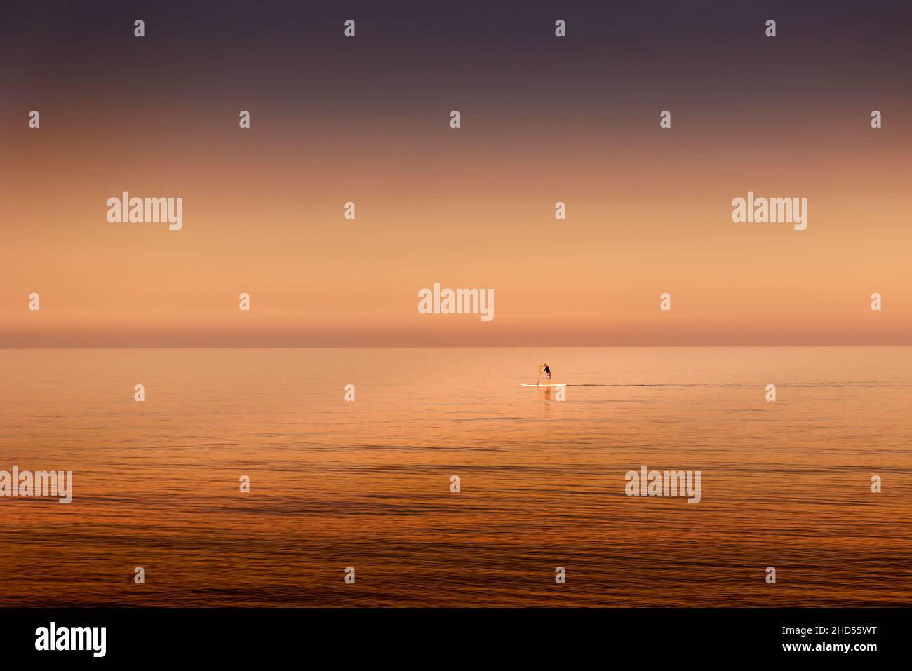 Surfer am Goldenen Meer. Landschaft in goldenen Tönen, kleine Figur eines Mannes im grenzenlosen Meer, ruhiger, offener Horizont. Stockfoto