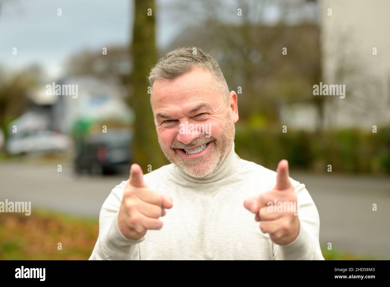 Fröhlich-beschwingter Mann grinst breit auf die Kamera, während er mit seinen Fingern auf die Kamera im Freien auf einer ruhigen Straße zeigt Stockfoto