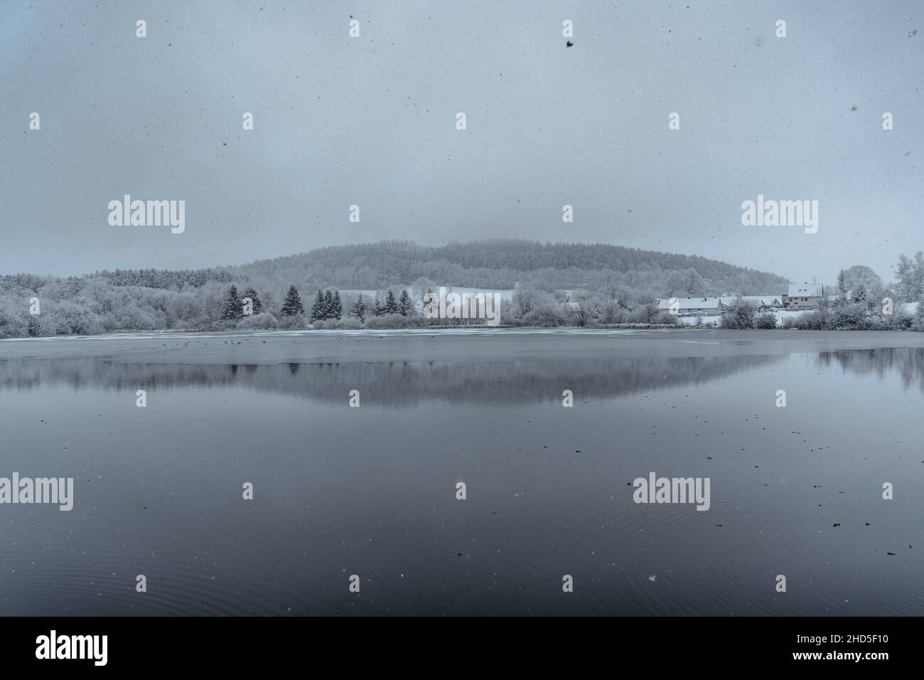 Bewölkt verschneiten Wintertag am See, kleines Dorf im Hintergrund, Bäume im Wasser reflektiert. Schneeflocken fallen. Teich in nebligen Landschaft. Kaltes Wetter, kein Peop Stockfoto