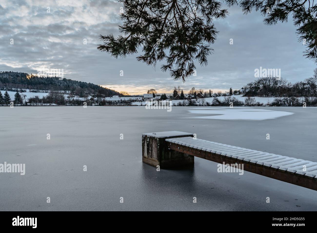 Hölzerner Pier am See mit frischem Schnee.Winterteich mit kleinem Steg bei Sonnenaufgang, Dorf im Hintergrund.Frostige ruhige Landschaft. Weiße Winterlandschaft verschneit Stockfoto