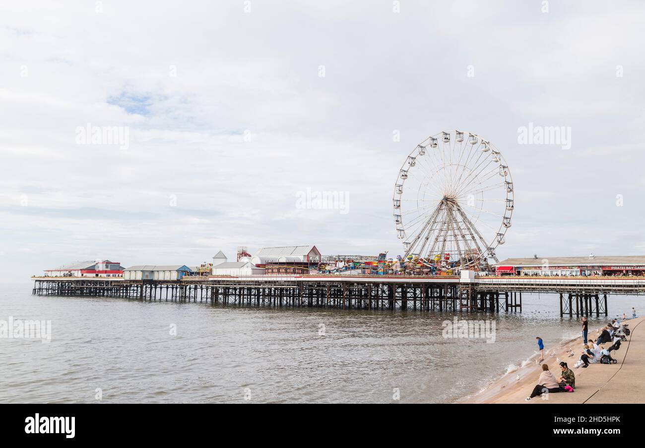 Touristen sitzen auf den Stufen am Strand von Blackpool und beobachten das Riesenrad, das sich am Central Pier dreht. Stockfoto