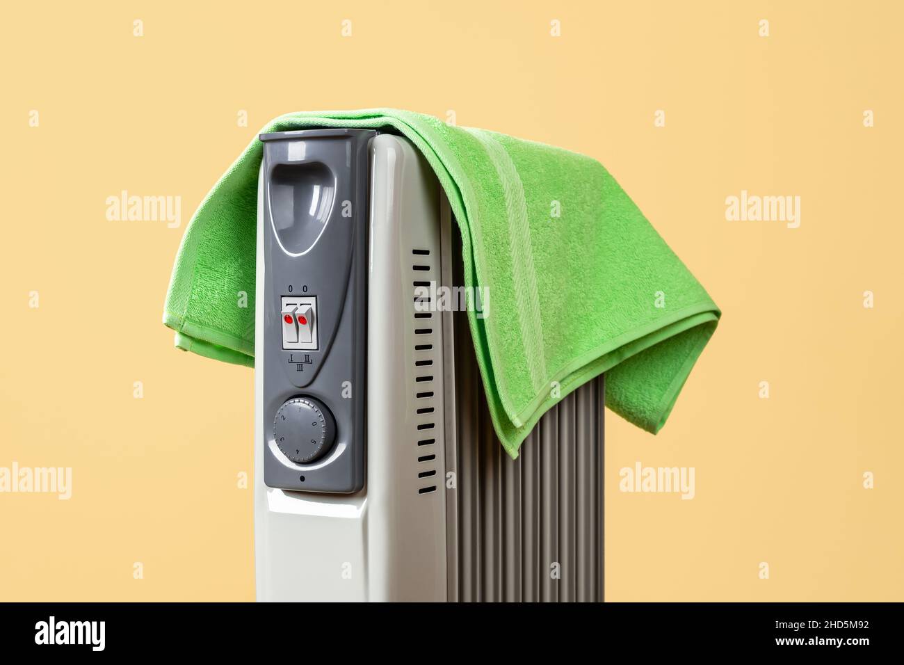 Grünes Handtuch trocknet auf dem mit heißem Öl gefüllten Heizgerät vor gelbem Hintergrund. Tragbare Raumheizung mit einem Handtuch bedeckt. Gefährliche Situation. Stockfoto