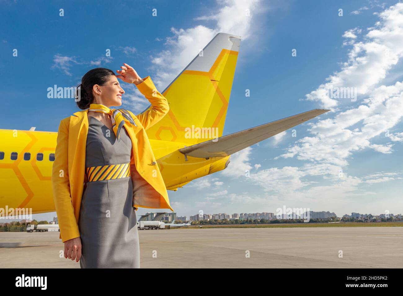 Fröhliche Flugbegleiterin in Flugzeuganzug, die mit der Hand die Augen vor der Sonne verhüllte und lächelte, während sie am bewölkten Himmel stand Stockfoto