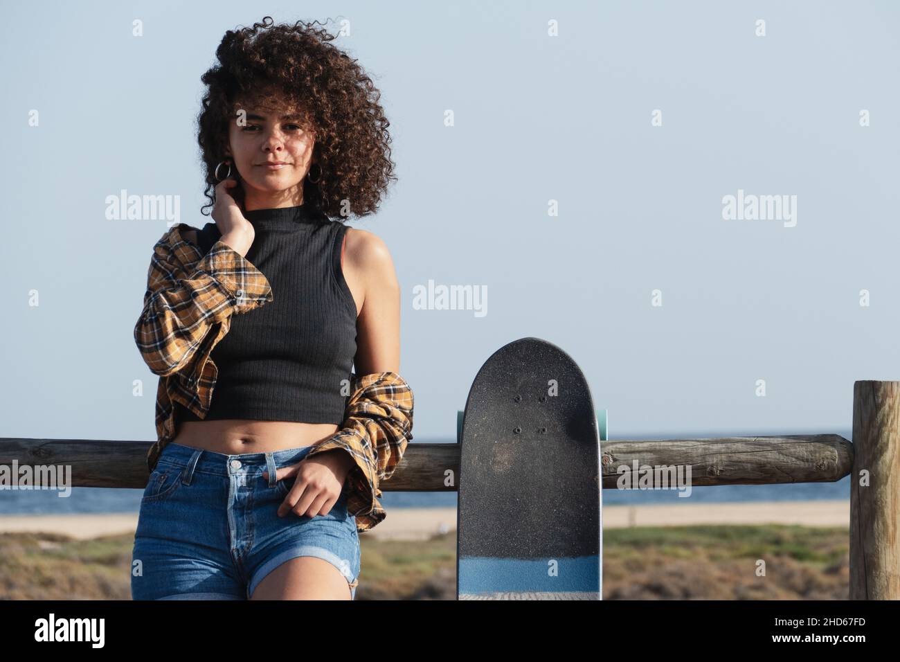 Hübsche junge Skaterin Frau mit Afro-Haar-Porträt. Sie ist im Freien, lehnt sich an einen Zaun und schaut auf die Kamera Stockfoto