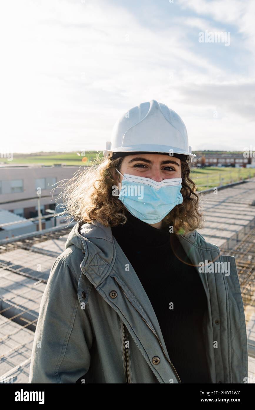 Lächelnde und fröhliche Architektin mit Hut und Gesichtsmaske, die auf einer Baustelle im Baumanagement arbeitet. Stockfoto