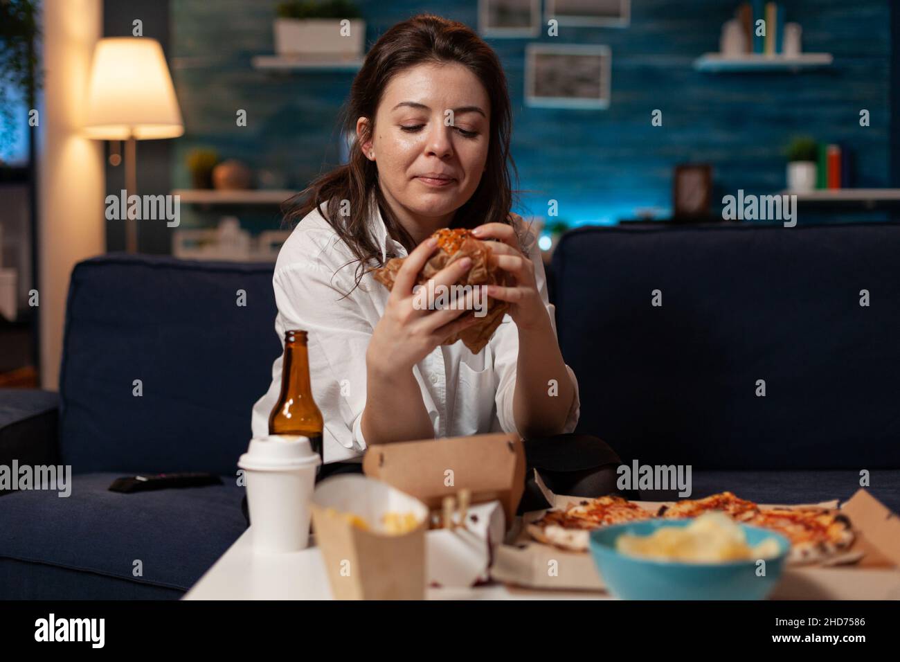 Frau, die auf der Couch sitzt und im Wohnzimmer vor dem Fernseher ein leckeres Abendessen mit Burger-Lieferung anschaut. Person, die am Tisch steht, mit einer großen Pizza zum Mitnehmen. Stockfoto