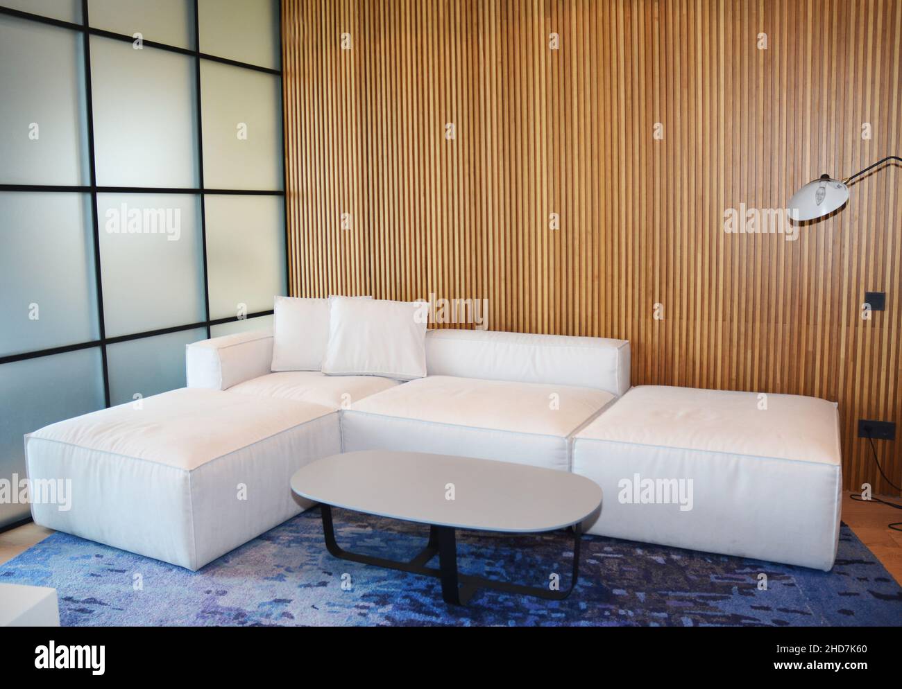 Ein Wohnzimmer modernes Innendesign mit einer Milchglaswand, Holzpaneelen, gemütlichem weißen Ecksofa mit einem ovalen Couchtisch auf einem blauen Teppich und mod Stockfoto