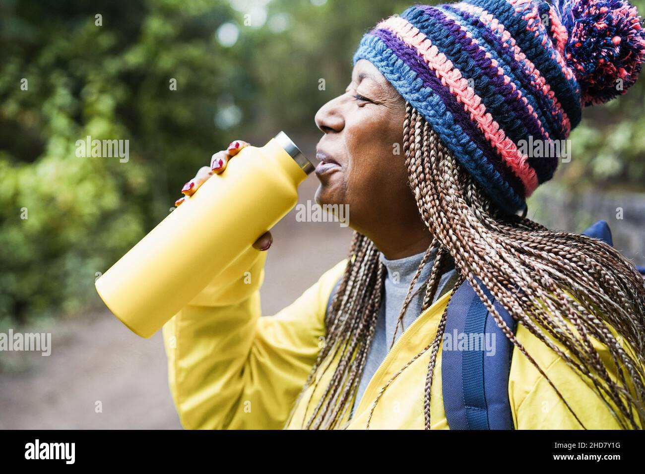 Ältere afrikanische Frau, die während des Trekkingtages im Bergwald Wasser trinkt - Fokus auf Hut Stockfoto