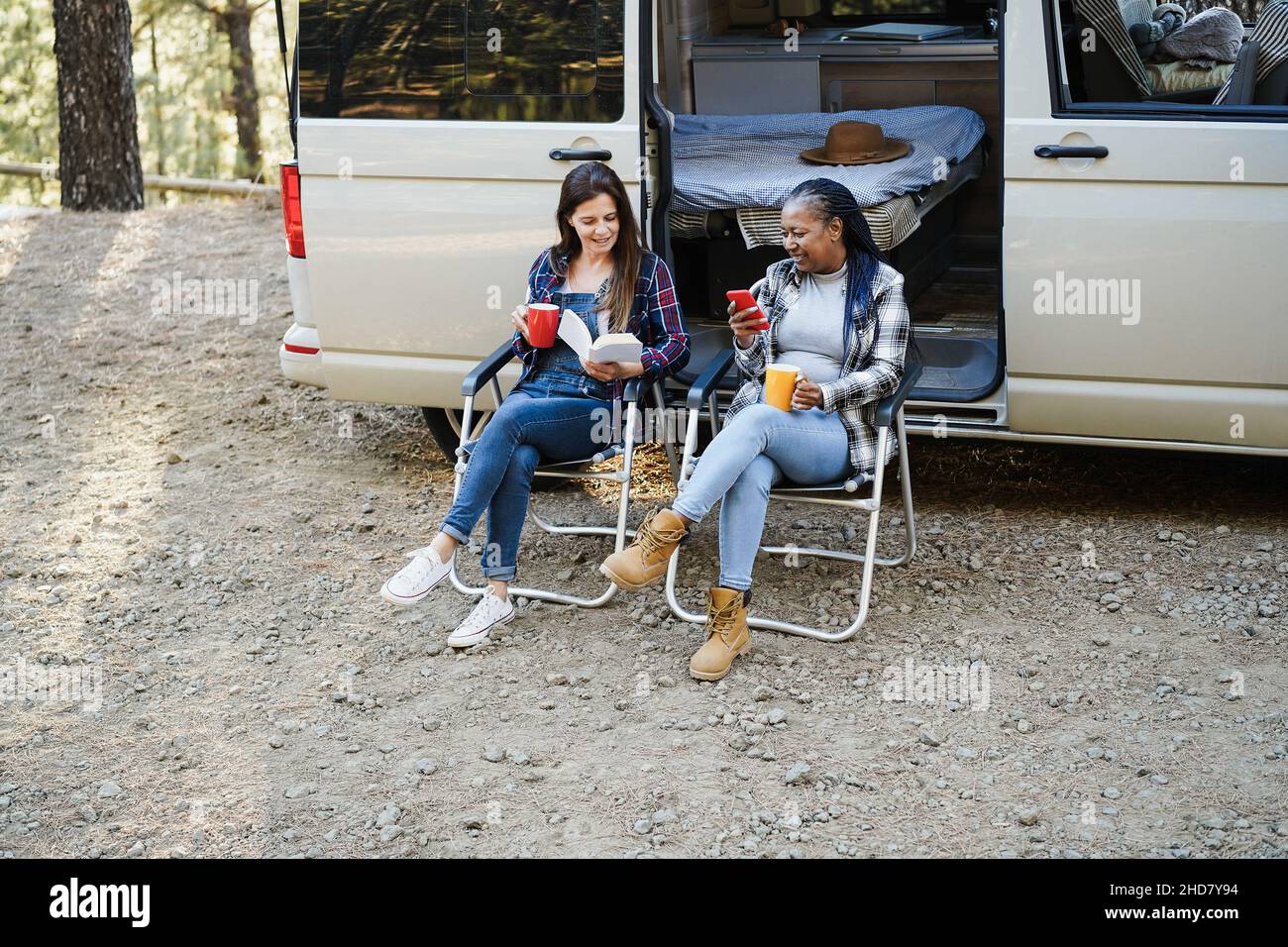 Multirassische Freundinnen, die Spaß beim Campen mit dem Wohnmobil haben, während sie im Freien Kaffee lesen und trinken - Fokus auf Gesichter Stockfoto