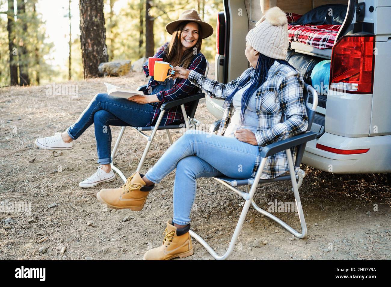Multirassische Freundinnen, die Spaß beim Campen mit Camper Van haben, jubeln mit Kaffee im Freien - Fokus auf Hand halten Tasse Stockfoto