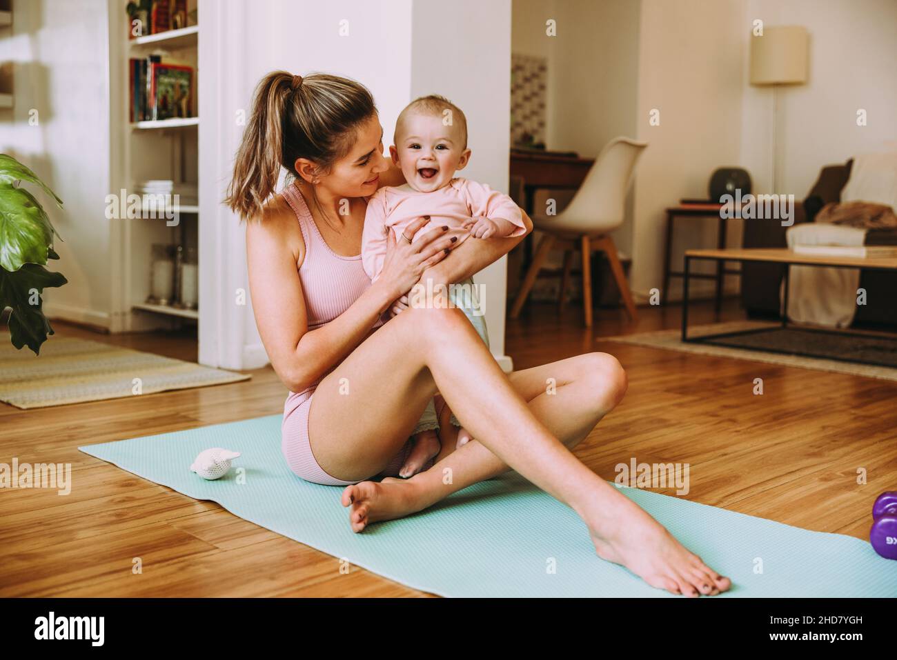 Entzückendes Baby, das aufgeregt in den Armen ihrer Mutter lächelt. Glückliche Mutter, die mit ihrem Baby auf einer Trainingsmatte trainiert. Liebevolle neue Mutter, die sich mit ihrem Baby verklebt Stockfoto